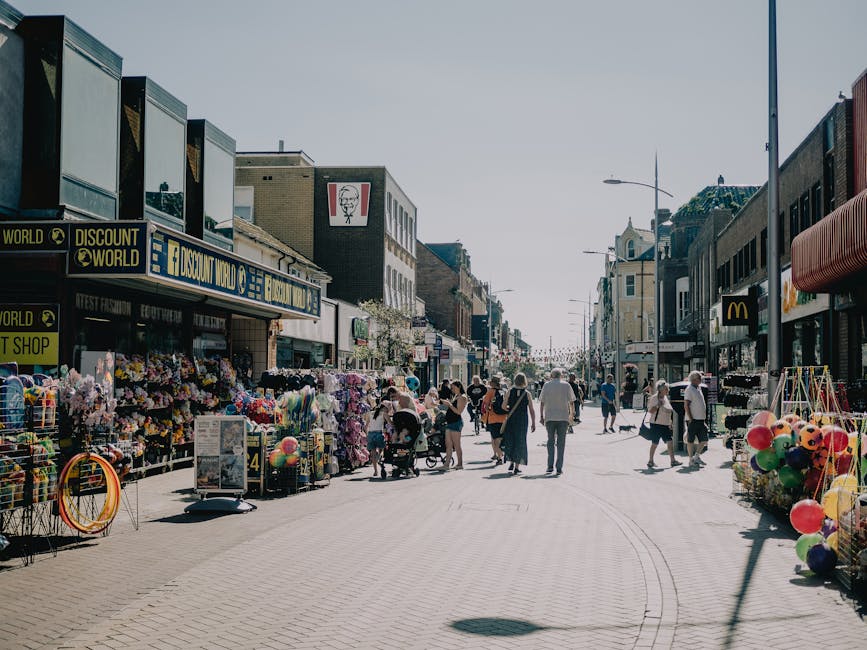 A busy street scene in a town centre during daytime, with pedestrians walking along a wide paved pedestrian area. On the left side, there are storefronts including a Discount World shop displaying col